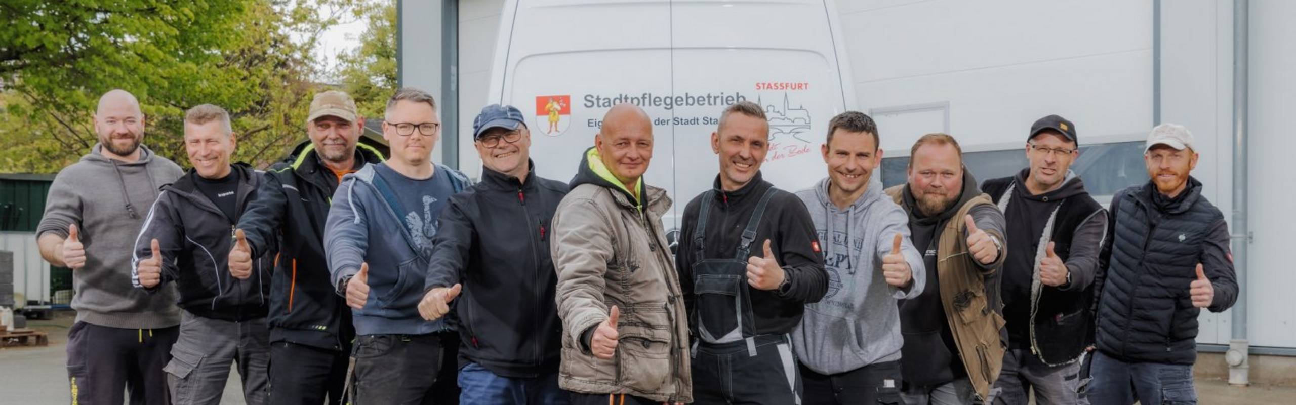 Elf Männer, die Hausmeister des Stadtpflegebetriebes, stehen nebeneinander vor einem Einsatzfahrzeug und zeigen die Daumen nach oben